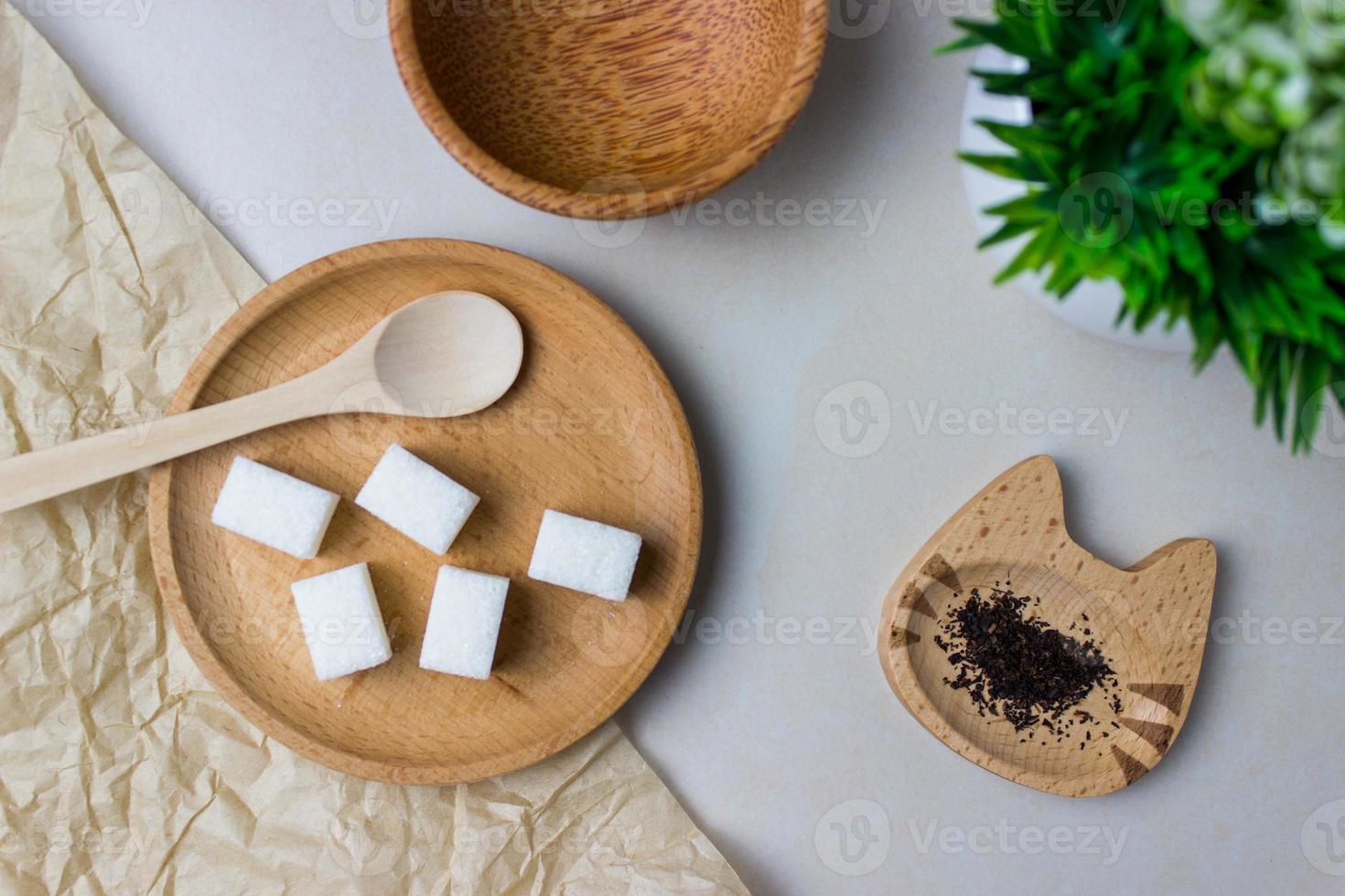 Sugar cubes in a wooden plate with spoon. Plate in the shape of a cat with tea feaves. On the kitchen table with green plant. Top view photo