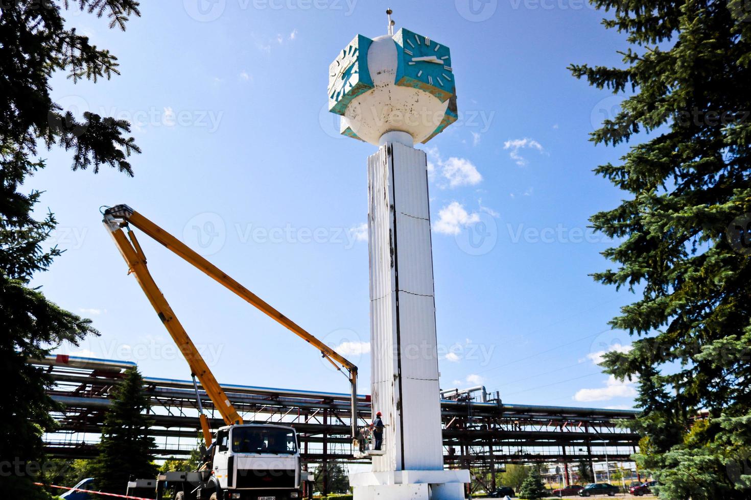 Repair of the tower clock. Workers on the crane repair clock-column, clock on the tower, paint, update, perform cosmetic repairs. Unusual clock against the blue sky photo
