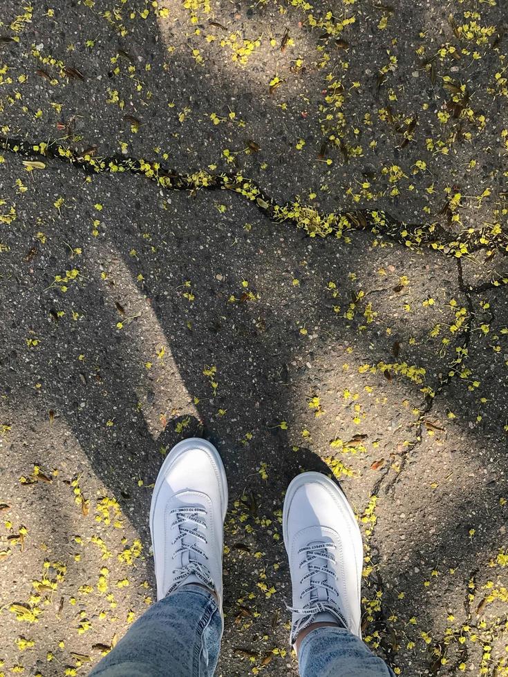 asphalt surface with uneven cracks through which small yellow flowers make their way. on the road are feet in fashionable white leather sneakers. there is a natural island in the city photo