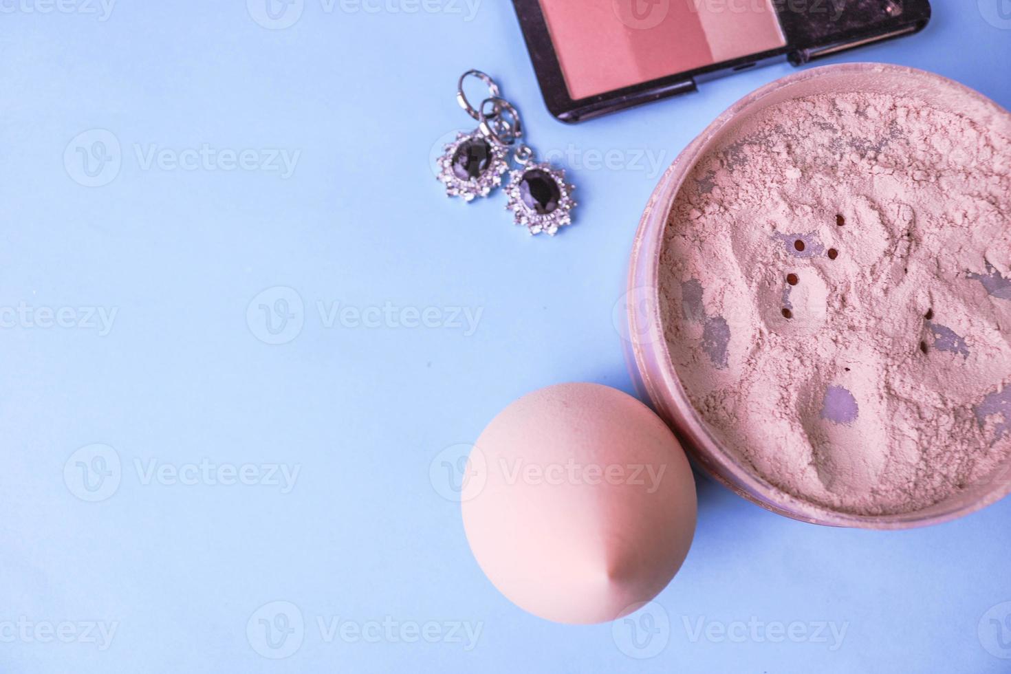 Beauty box, loose matte mineral powder and blush with a beauty blender for makeup and silver earrings on a blue background. Flat lay. Top view photo