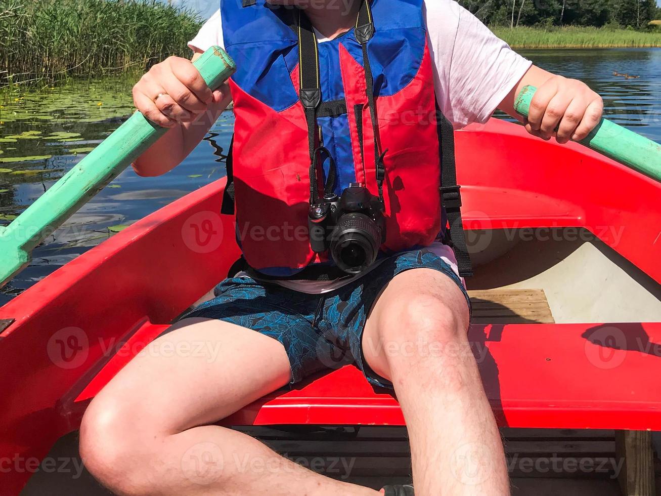 A man in a red boat, shorts and a life jacket is paddling with oars on a boat for a walk on the water of the lake river sea in nature photo