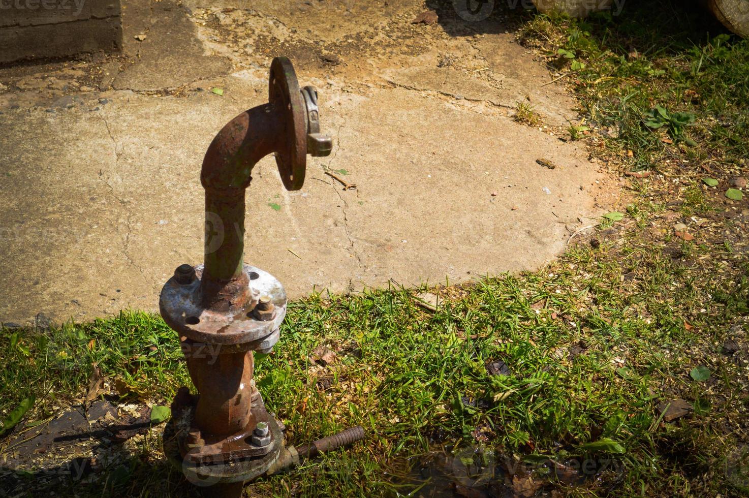 una vieja tubería de suministro de agua industrial oxidada y corrosiva de hierro para el suministro de agua con una brida que se mueve con tuercas y espárragos sobresale del suelo de hierba verde foto