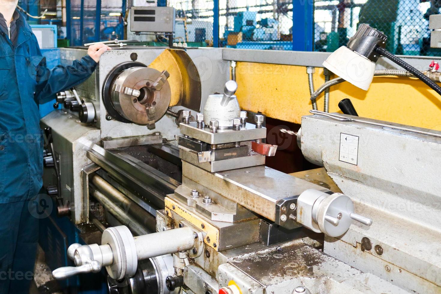 un hombre que trabaja con una túnica, overol se encuentra junto a un torno industrial para cortar, tornear cuchillos de metales, madera y otros materiales, tornear, hacer detalles y repuestos en la fábrica foto