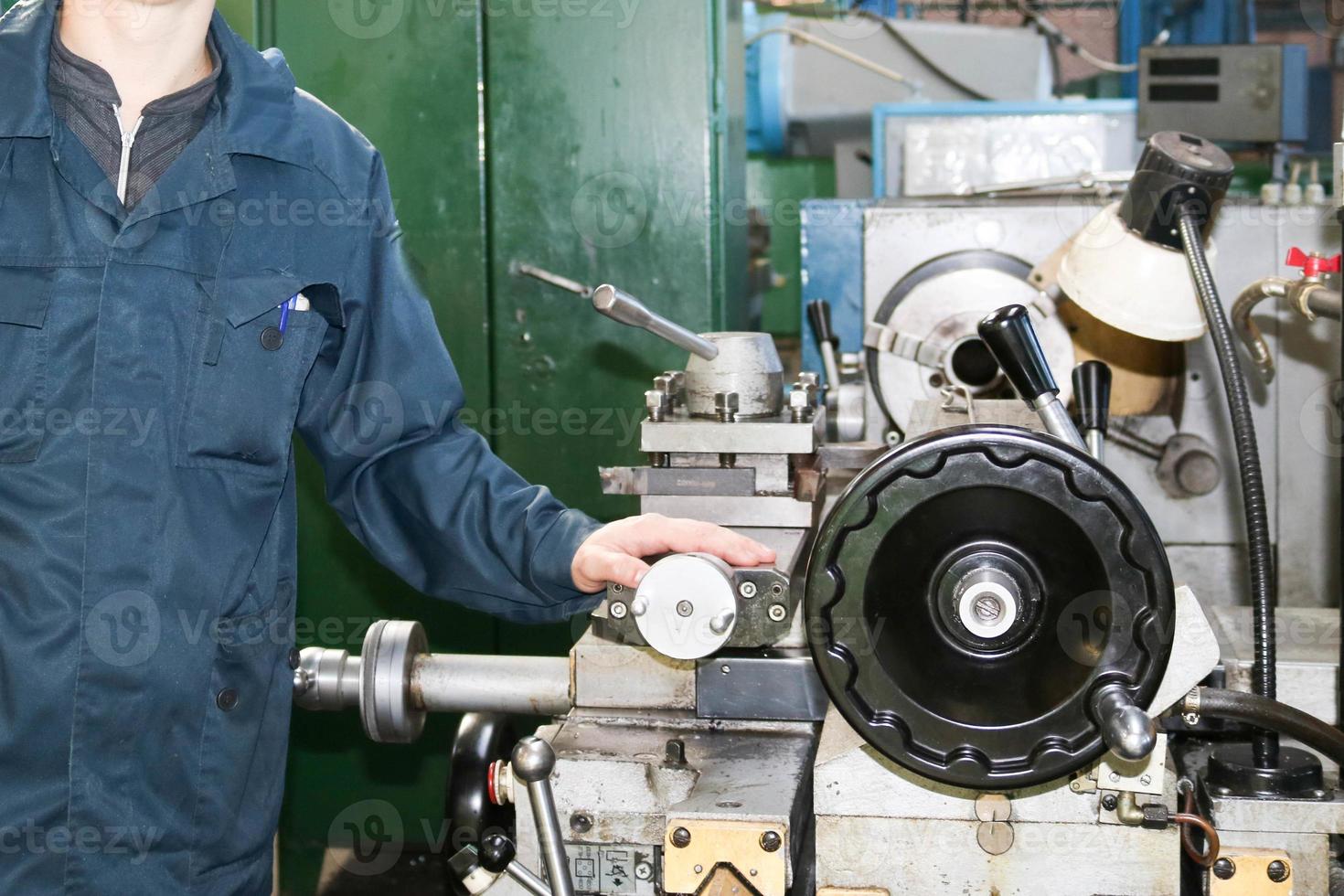 un hombre que trabaja con una túnica, overol se encuentra junto a un torno industrial para cortar, tornear cuchillos de metales, madera y otros materiales, tornear, hacer detalles y repuestos en la fábrica foto