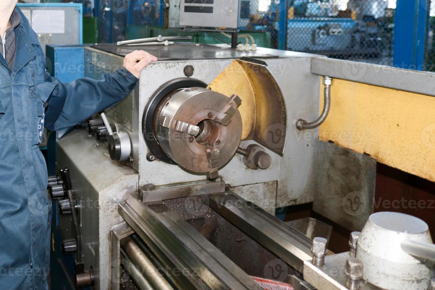 un hombre que trabaja con una túnica, overol se encuentra junto a un torno industrial para cortar, tornear cuchillos de metales, madera y otros materiales, tornear, hacer detalles y repuestos en la fábrica foto