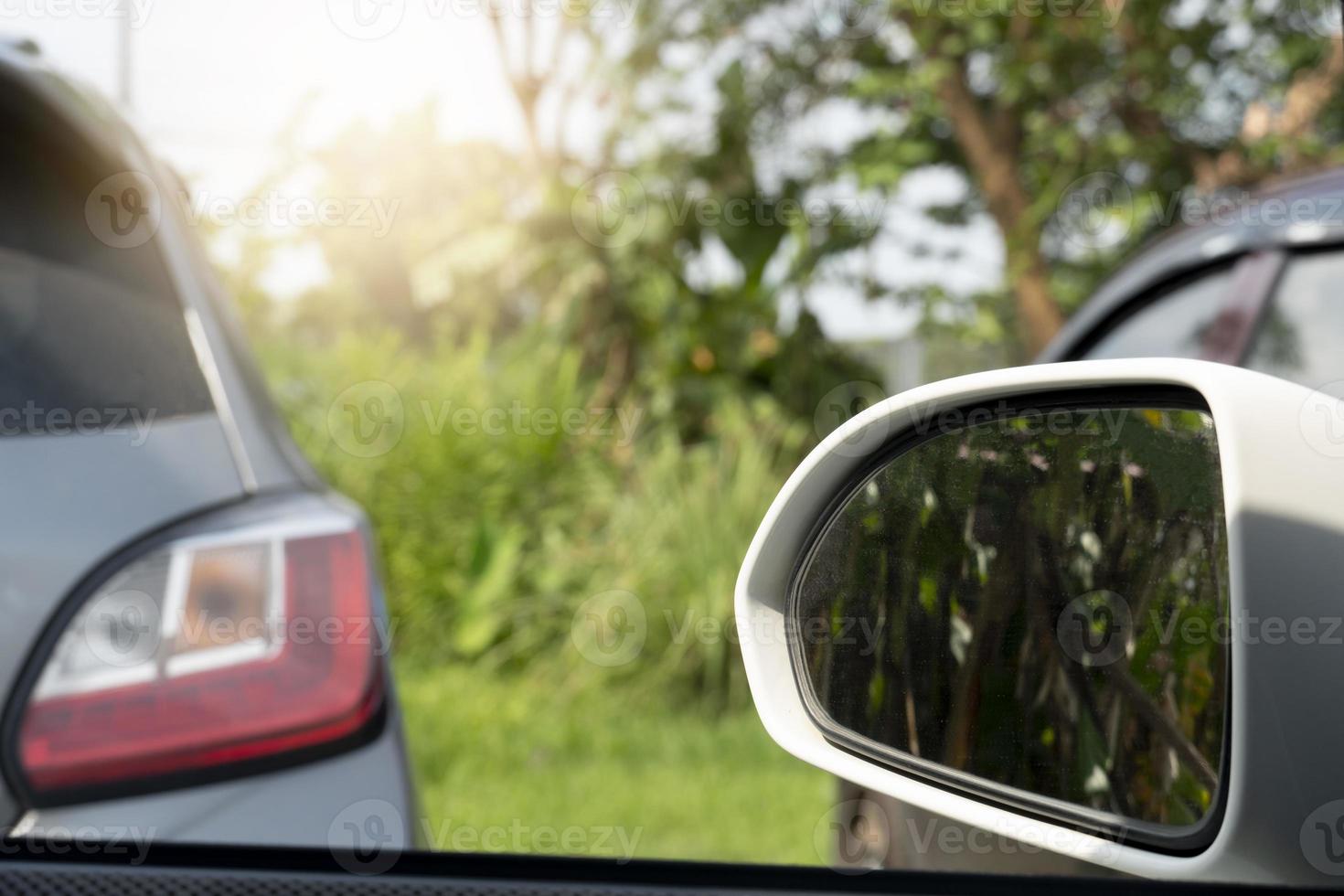Mirror wing of white car driving on the road. Pass through other car in the parking lot. Parking area with green grass and trees. photo