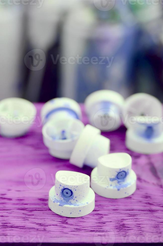 The soiled blue nozzles from the paint sprayer lies on a purple plank on a background of a many dirty spray cans photo