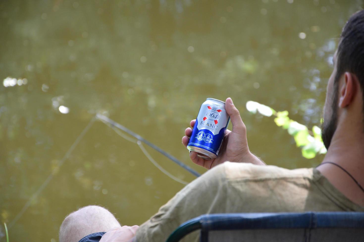 KHARKIV, UKRAINE - MAY 02, 2021 Young man holds 1664 Kronenbourg Blanc beer can outdoors during fishing. 1664 Blanc is the wheat beer from the French brewery Kronenbourg exported worldwide photo