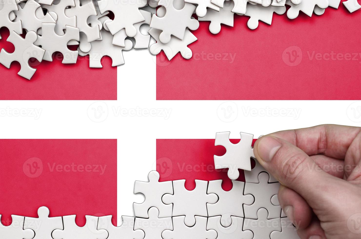 Denmark flag  is depicted on a table on which the human hand folds a puzzle of white color photo