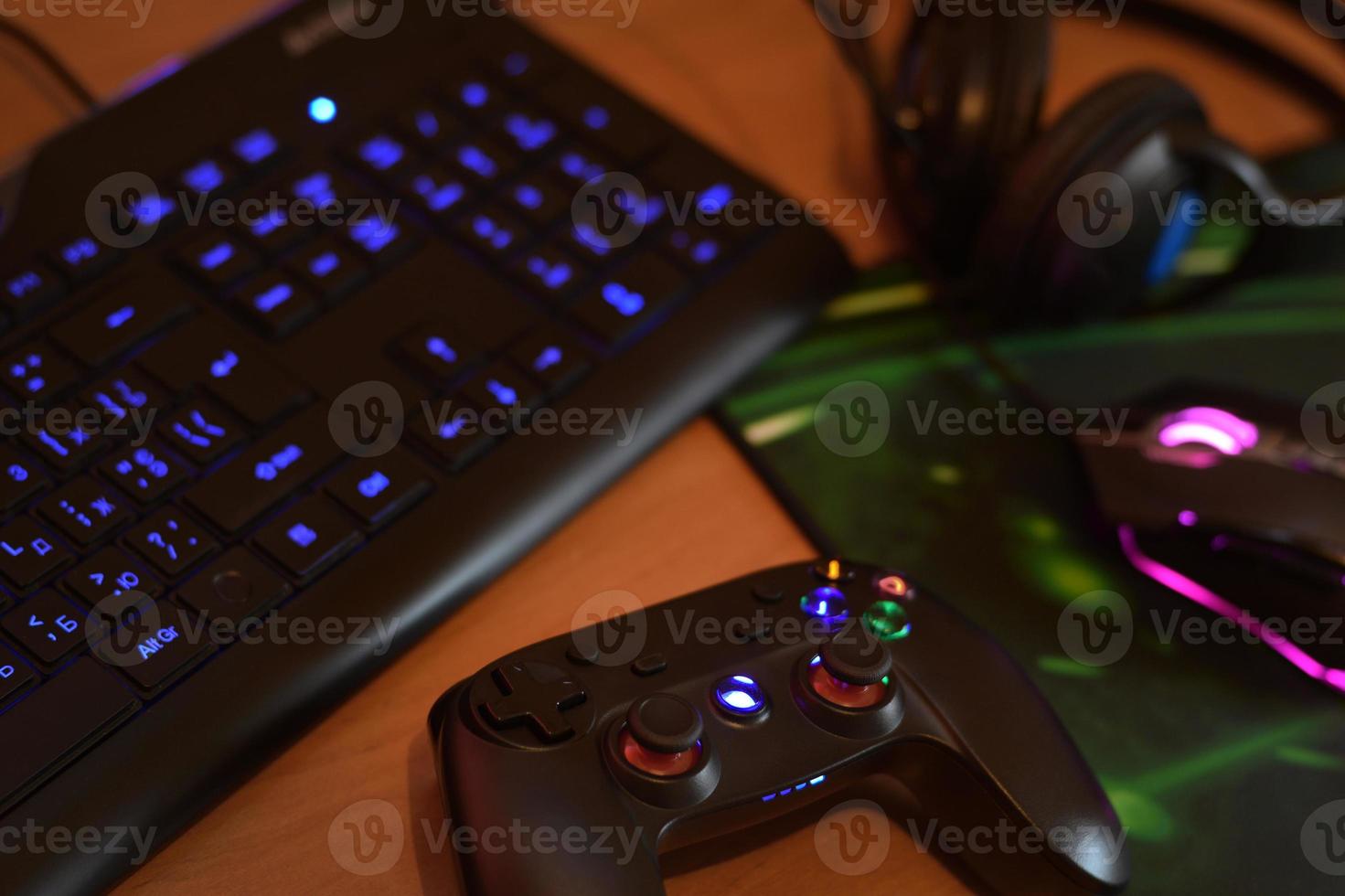 Modern gamepad and gaming mouse lies with keyboard and headphones on table in dark playroom scene. Modern gaming electronics shop concept photo