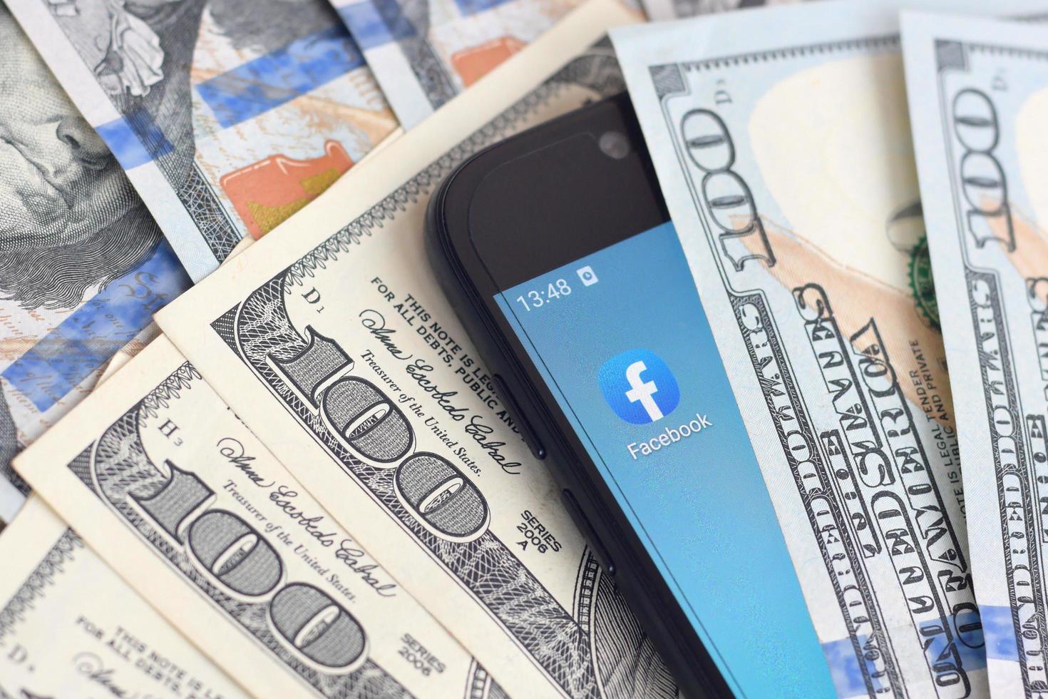 KHARKIV, UKRAINE - MAY 2, 2021 Smartphone screen with Facebook app and lot of hundred dollar bills. Business and social networking concept photo