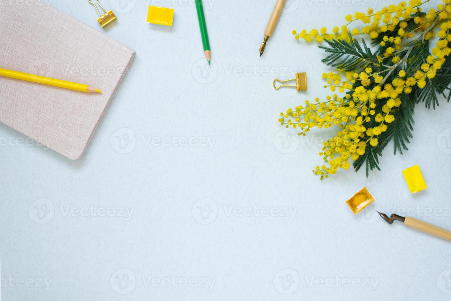 A frame of notepads, colored pencils, watercolors, mimosa branches and a pen on a blue background. Copy space for a blogger, artist, or calligrapher photo