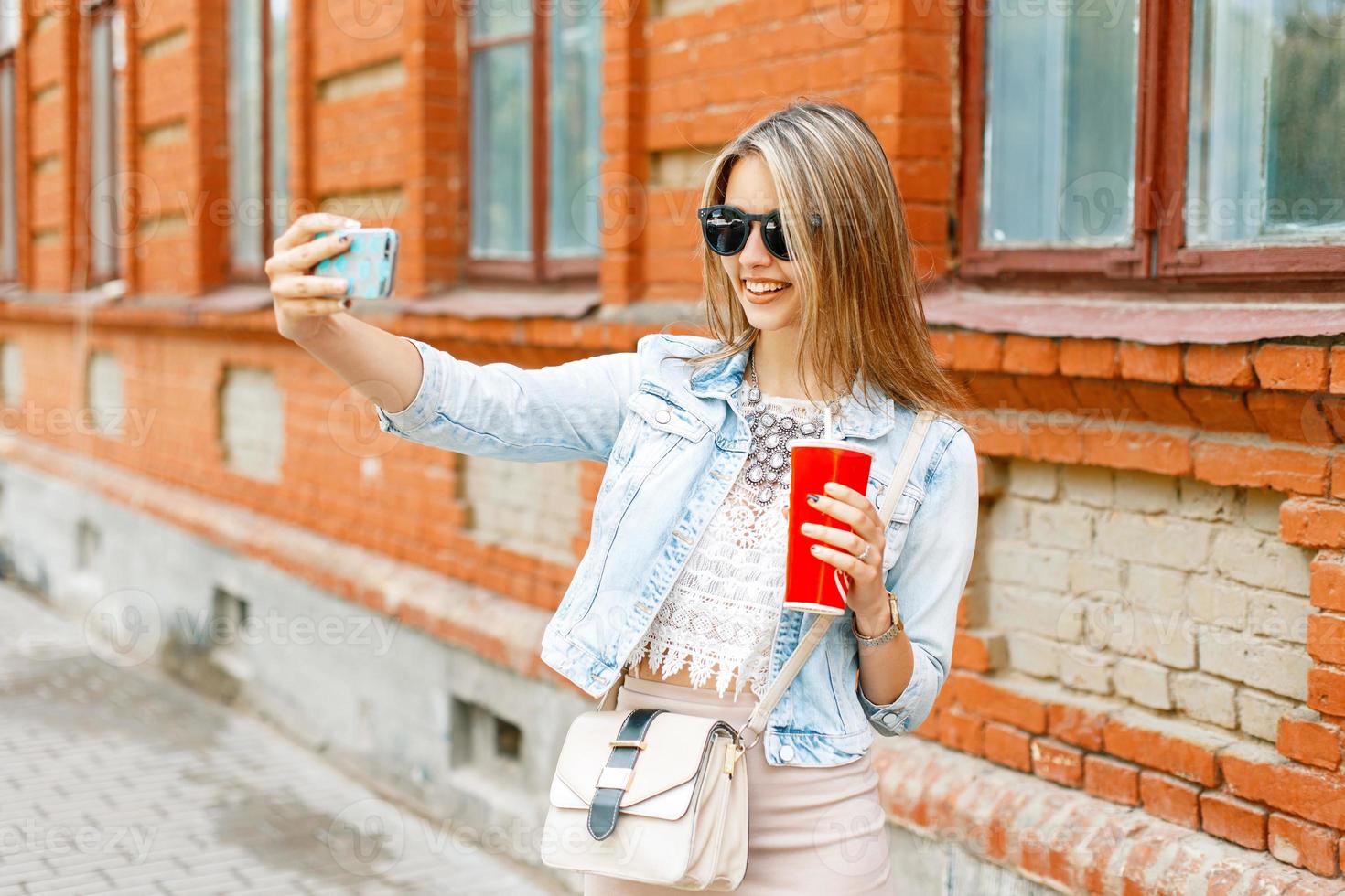 hermosa mujer alegre con una sonrisa en una chaqueta de denim sosteniendo una taza roja con bebida y fotografiada por teléfono en un día de verano. foto