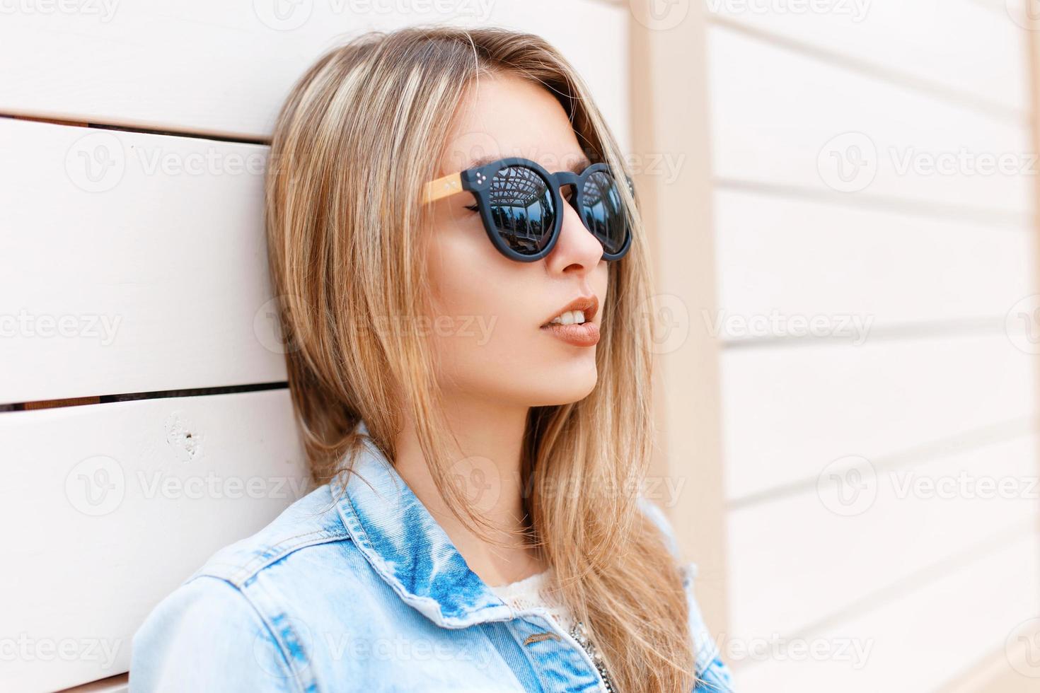 retrato de primer plano de una hermosa joven con gafas de sol y chaqueta vaquera en la playa cerca de la pared de madera foto