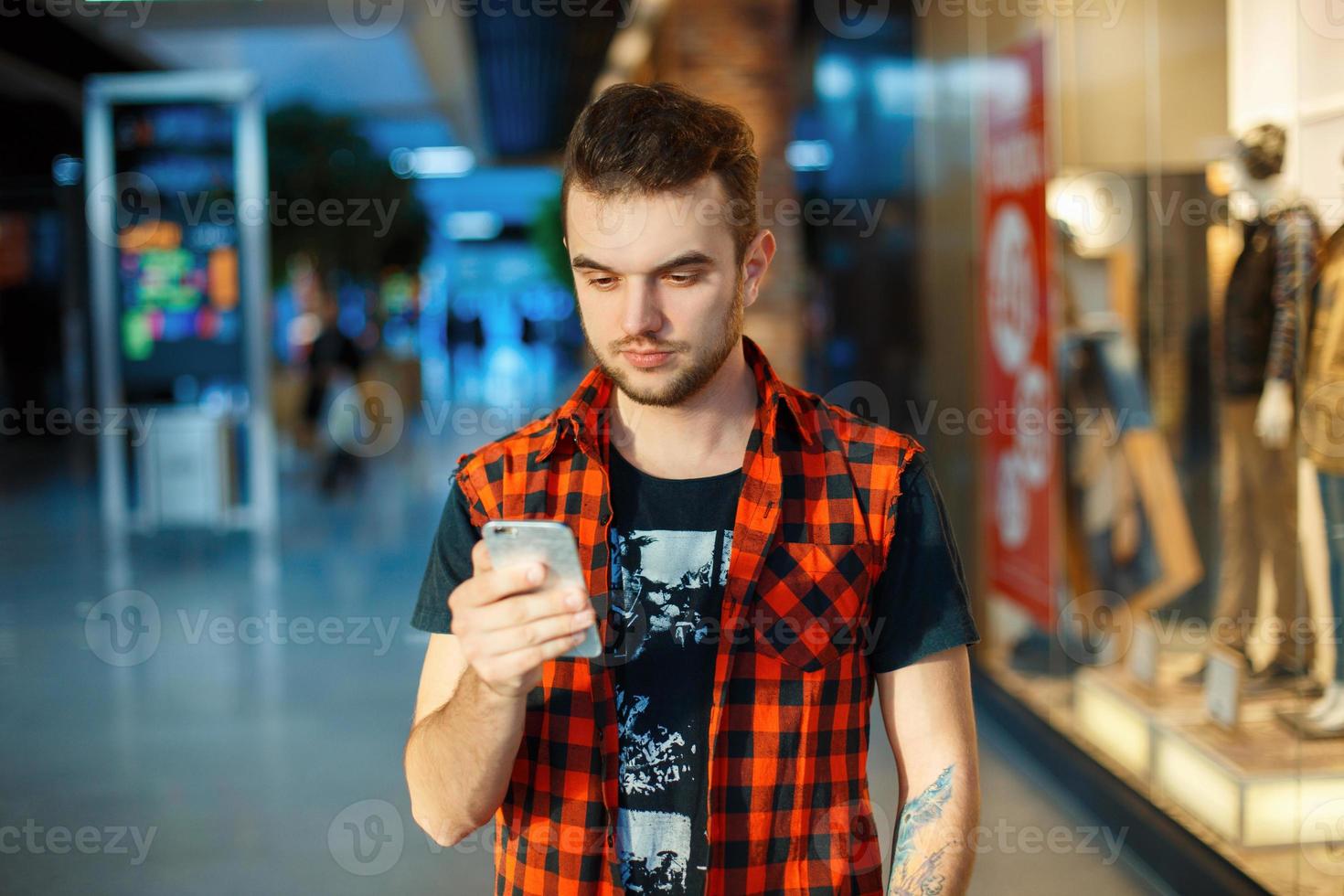 Young handsome man in the red shirt is holding a phone on the background of a shopping center photo