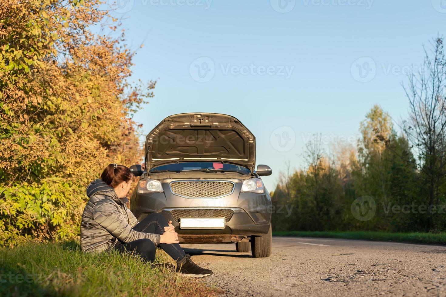 A woman in a jacket is trying to fix a broken car on the road. photo