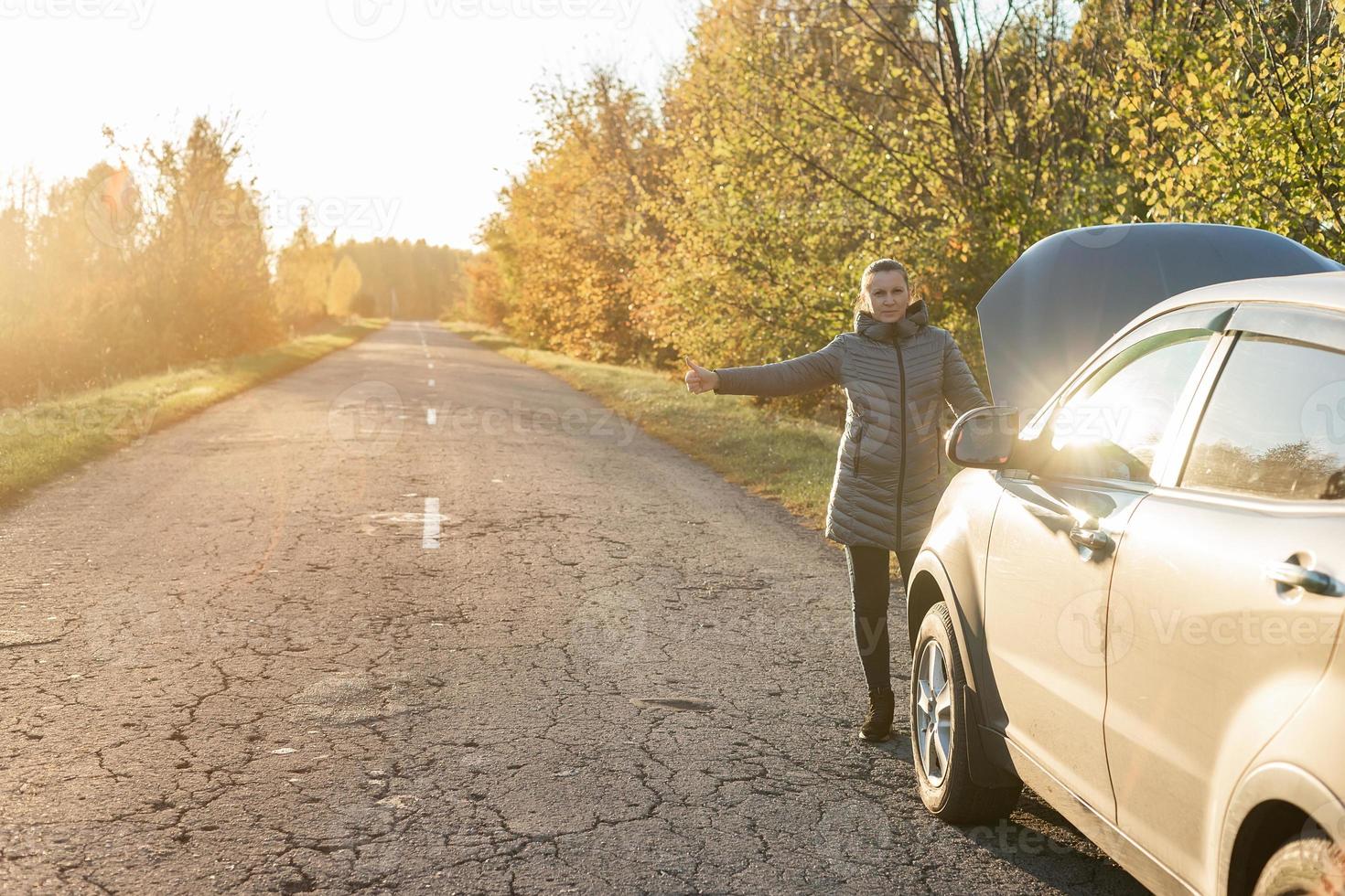 A woman in a jacket is trying to fix a broken car on the road. photo