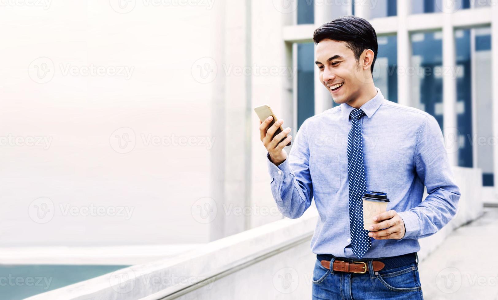 Smiling Young Asian Businessman Using Mobile Phone in the City. Modern Urban Lifestyle. Male Portrait. Hand holding Coffee Cup. looking at Smartphone photo