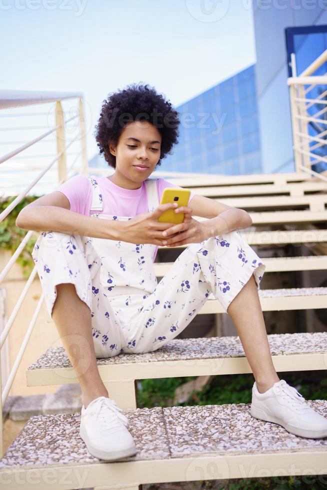 Black woman texting with her smartphone, sitting on steps of a modern building. photo