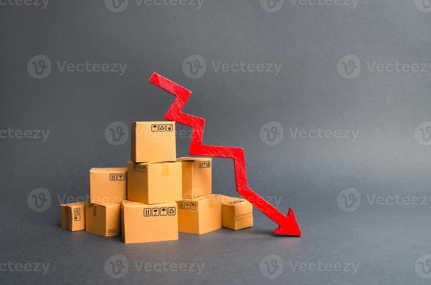 A pile of cardboard boxes and a red arrow down. The decline in the production of goods and products, the economic downturn and recession. Falling consumer demand, declining exports or imports. photo