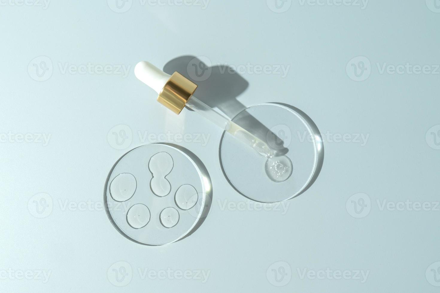 A transparent face serum or oil in a dropper bottle with drops on a round podiums. Cosmetology and pharmaceuticals concept. photo