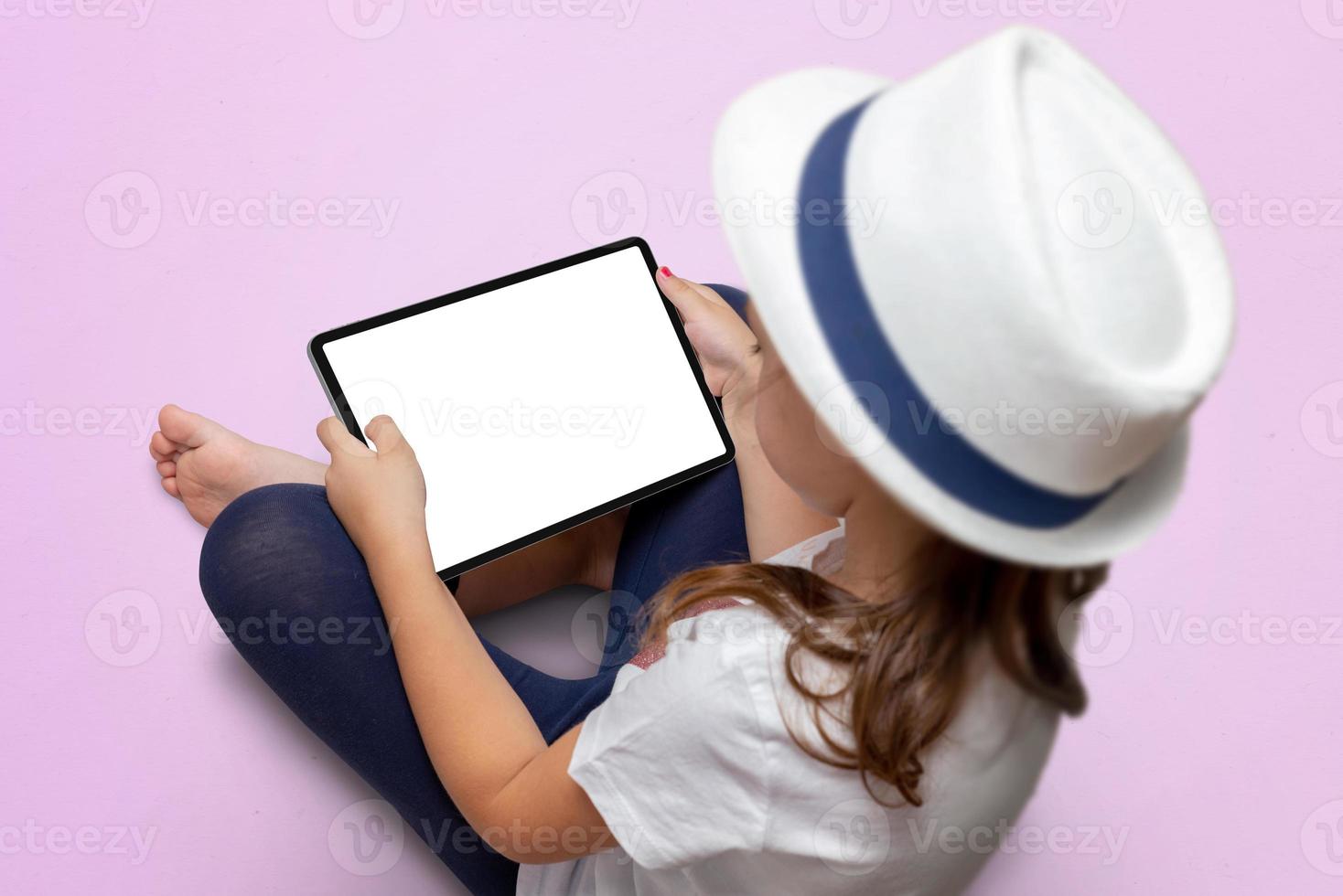 Little girl with a white hat is sitting on a pink floor and holding a tablet with isolated screen. Tablet mockup photo