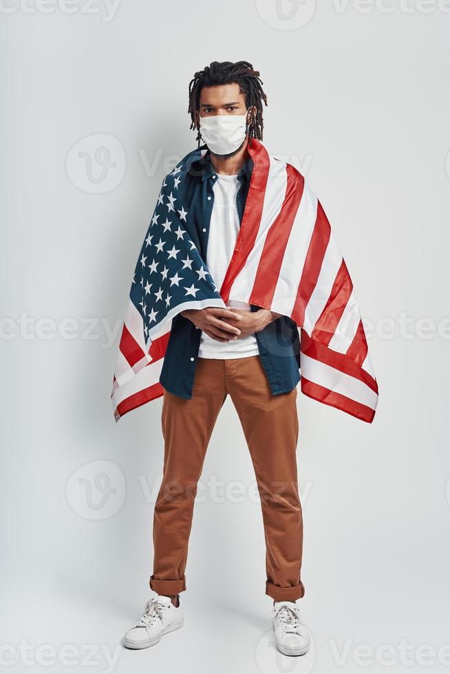 Full length of handsome young African man covered with American flag wearing medical face mask and looking at camera while standing against grey background photo