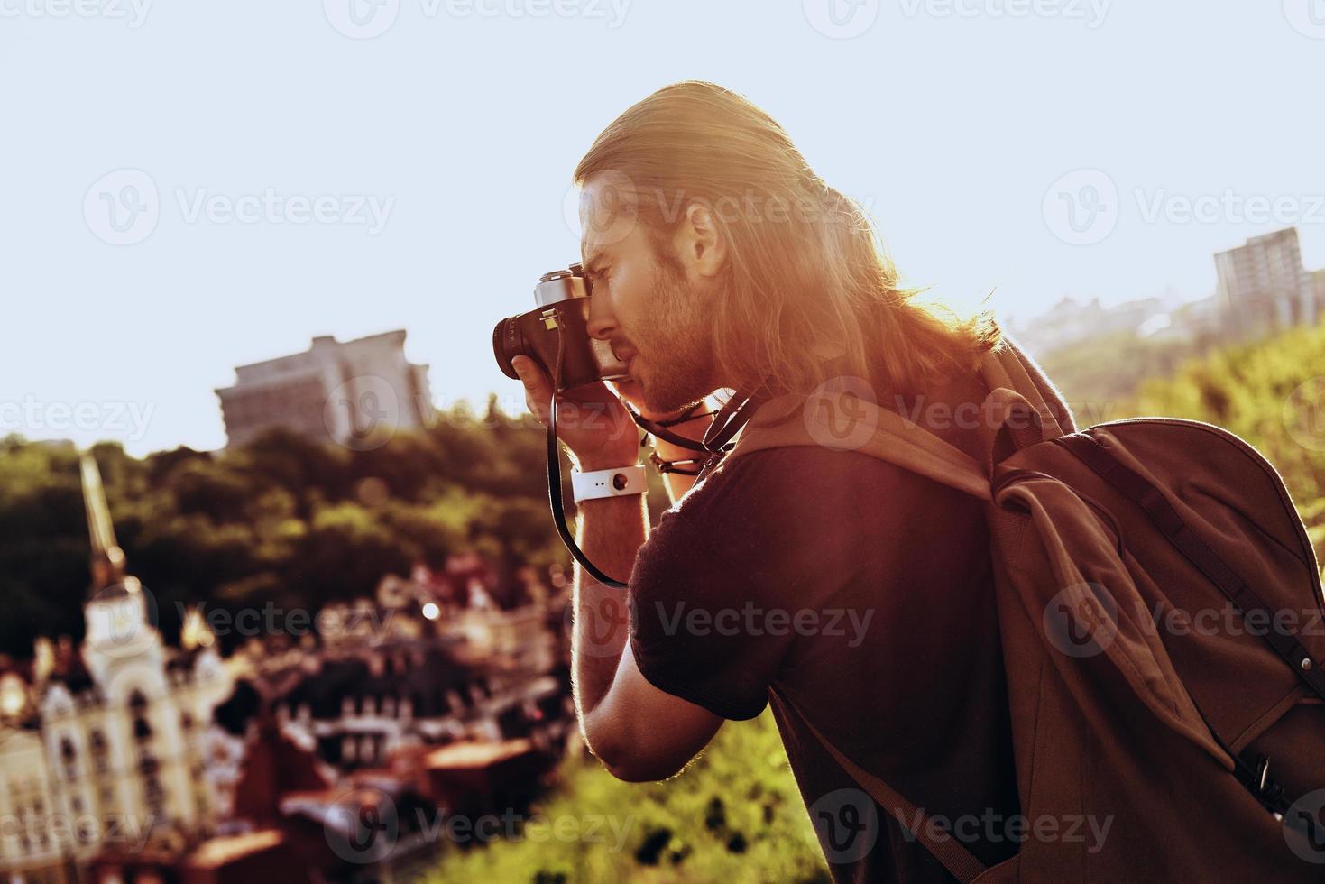 Nice shot. Young man in casual clothing photographing the view while standing on the hill outdoors photo