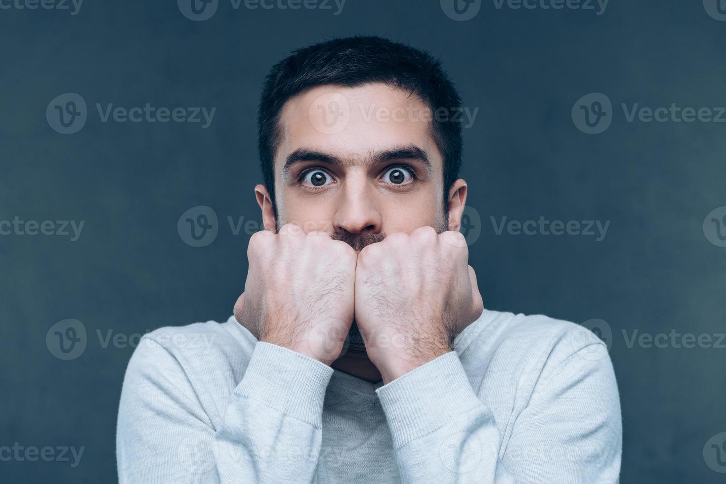 Feeling shocked. Terrified young man looking at camera and biting nails while standing against grey background photo