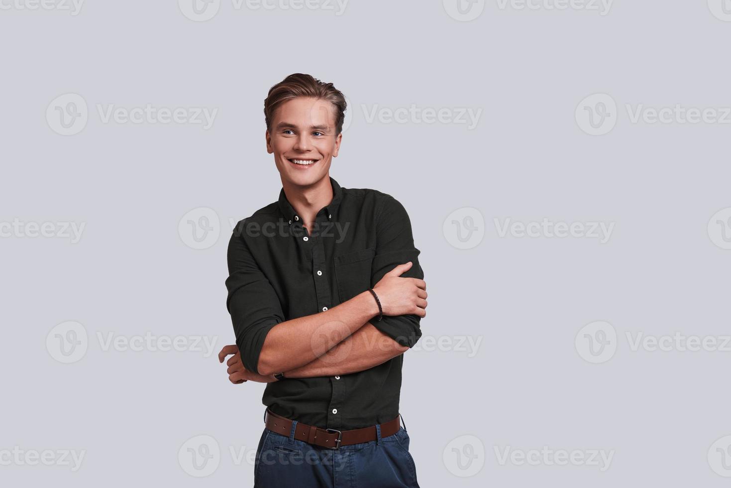 Young and confident. Good looking young man keeping arms crossed and looking away while standing against grey background photo