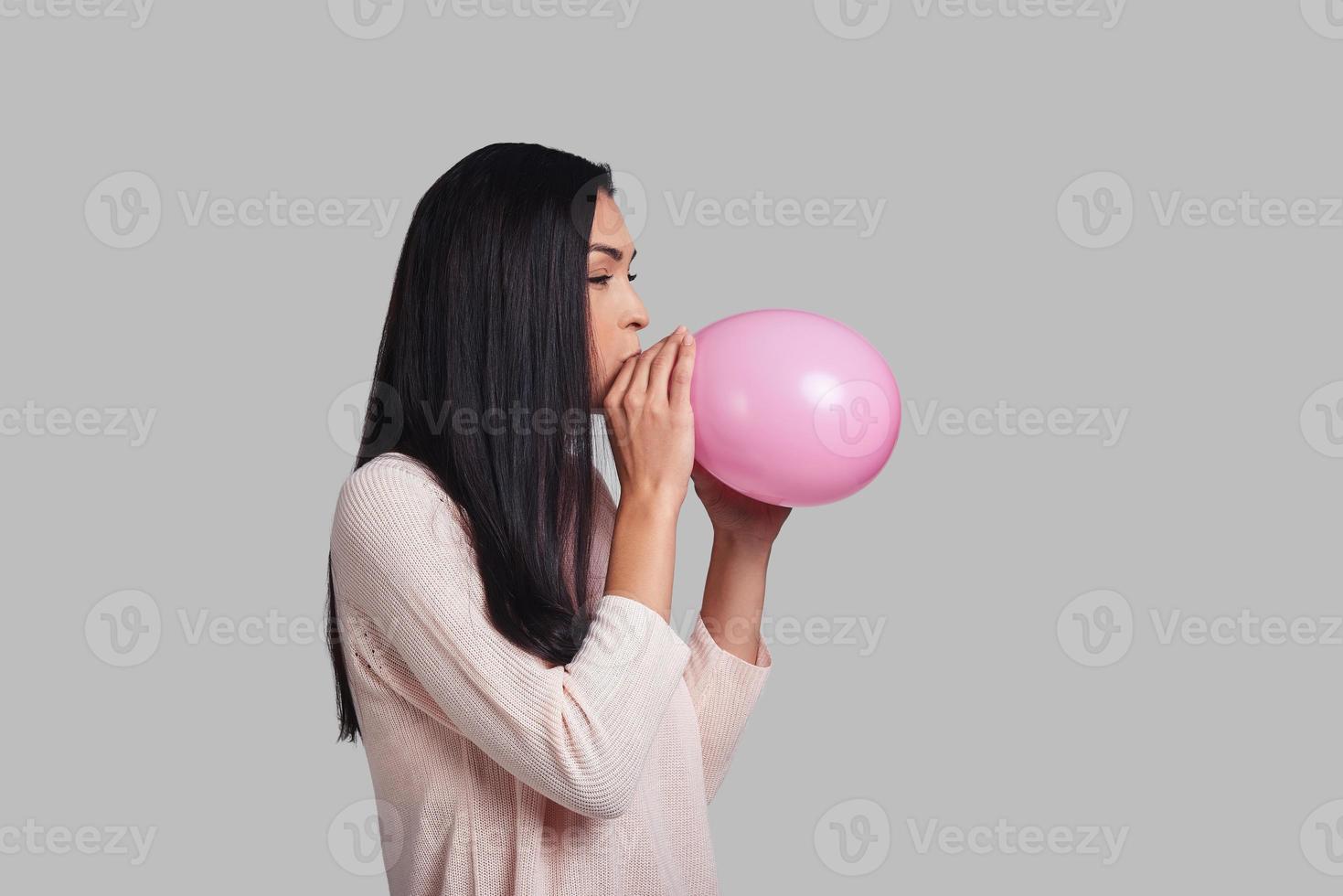 In a mood for a big holiday. Studio shot of attractive young woman in casual wear blowing up a pink balloon while standing against grey background photo