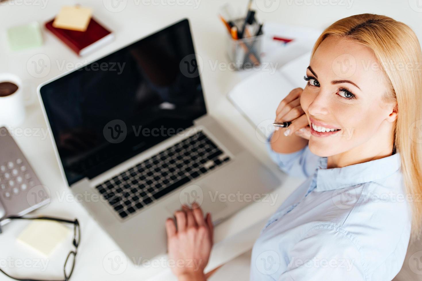 Beauty at work. Top view of cheerful young beautiful woman working on laptop and looking at camera with smile while sitting at her working place photo