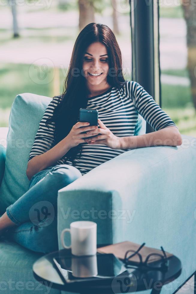 Sending message to him. Beautiful young smiling woman looking at her smart phone while sitting in a big comfortable chair at home photo