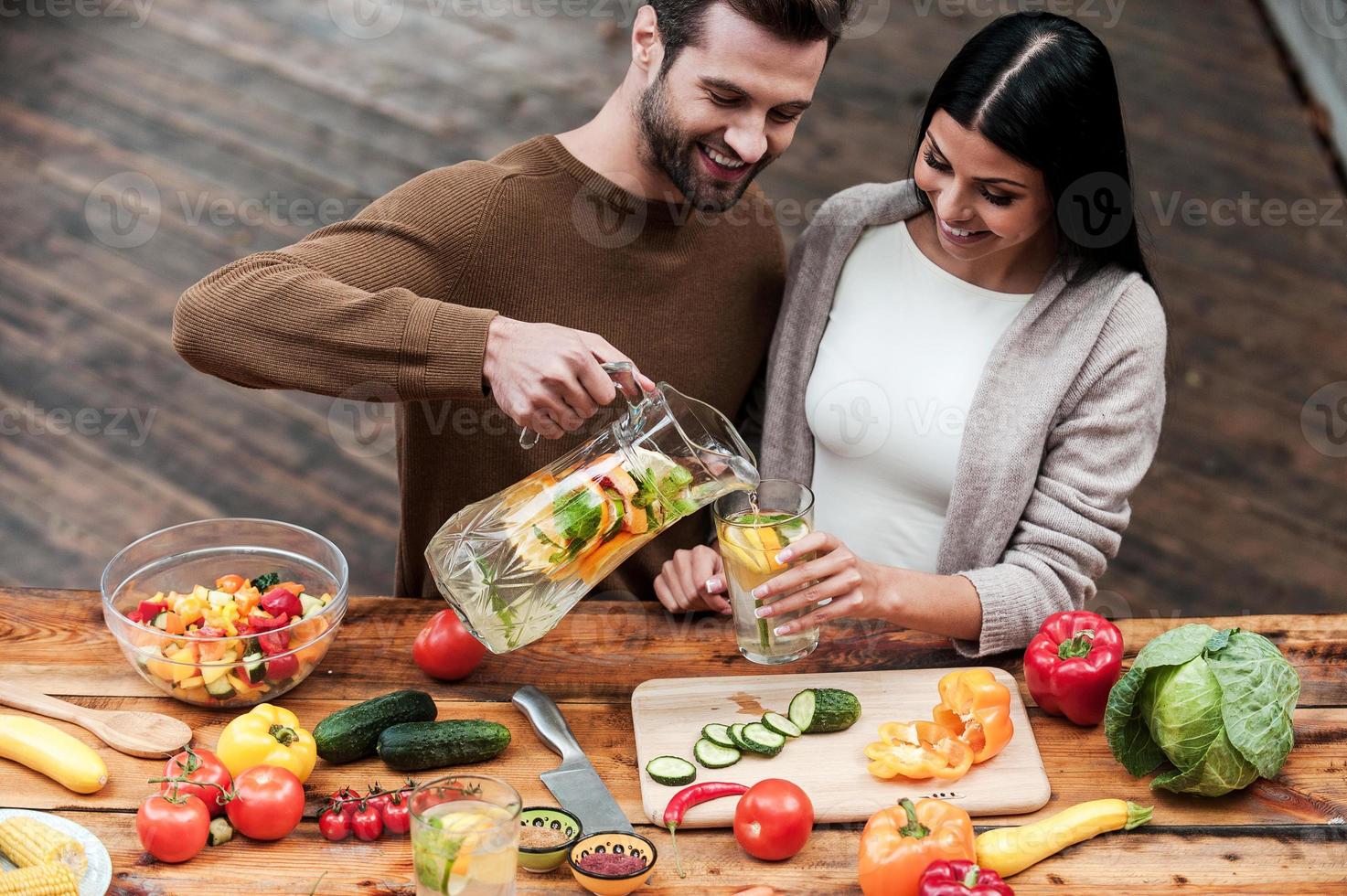 Enjoying healthy food and drinks. Cheerful young man pouring fresh lemonade to his girlfriend while preparing food on the wooden desk together photo