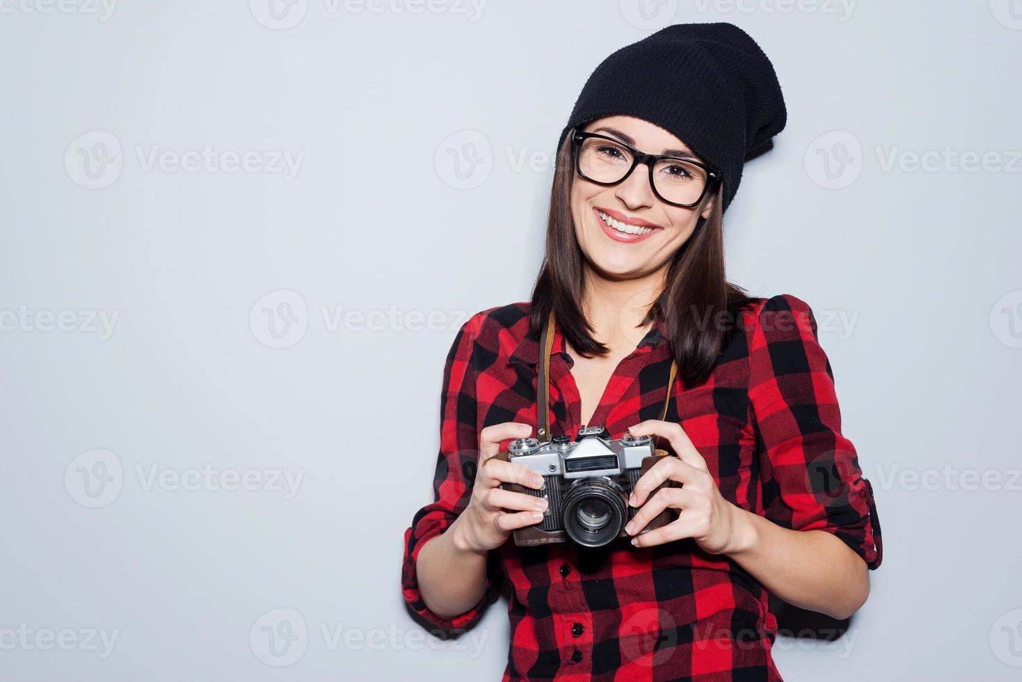 Stylish photographer.  Beautiful young woman in headwear and glasses holding camera and looking at camera while standing against grey background photo