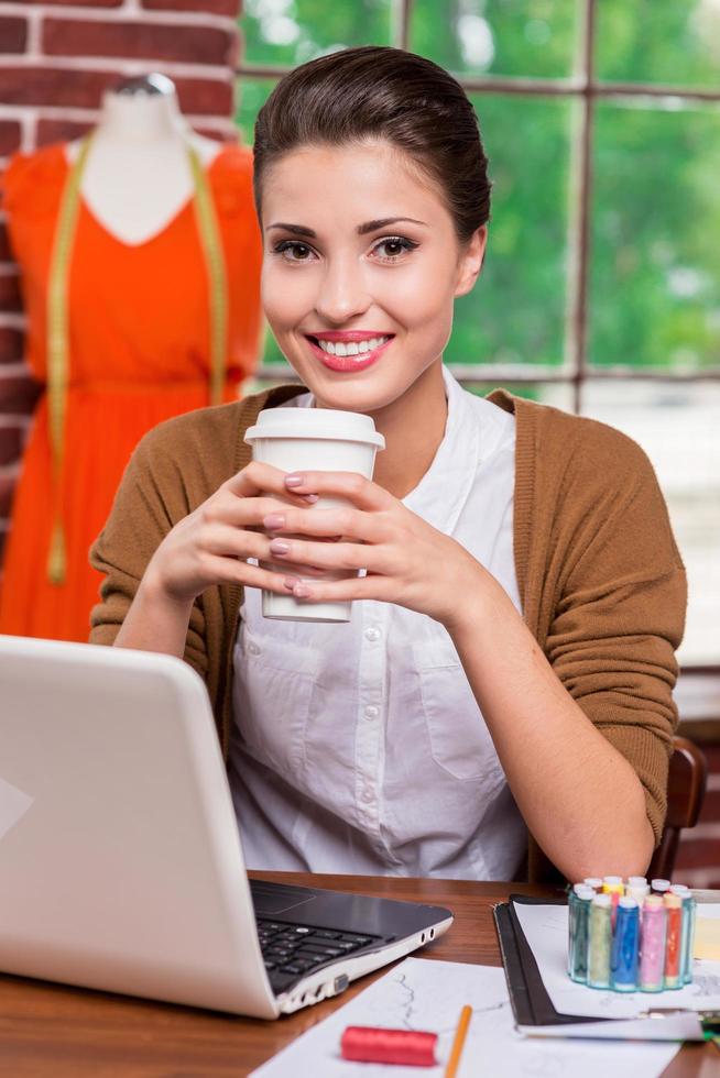 Refreshing her mind. Cheerful young fashion designer holding cup with hot drink and smiling while sitting at her working place and with mannequin standing in the background photo