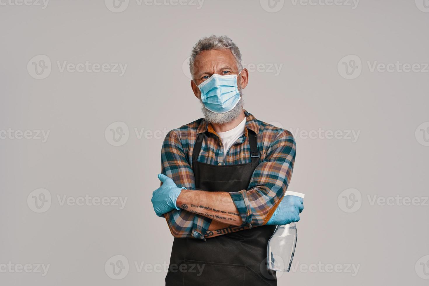 Senior man in apron and protective face mask looking at camera while standing against gray background photo