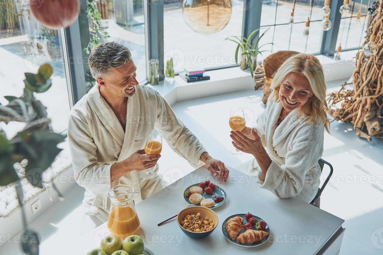 vista superior de una hermosa pareja madura en albornoces disfrutando del desayuno juntos mientras pasan tiempo en la cocina doméstica foto