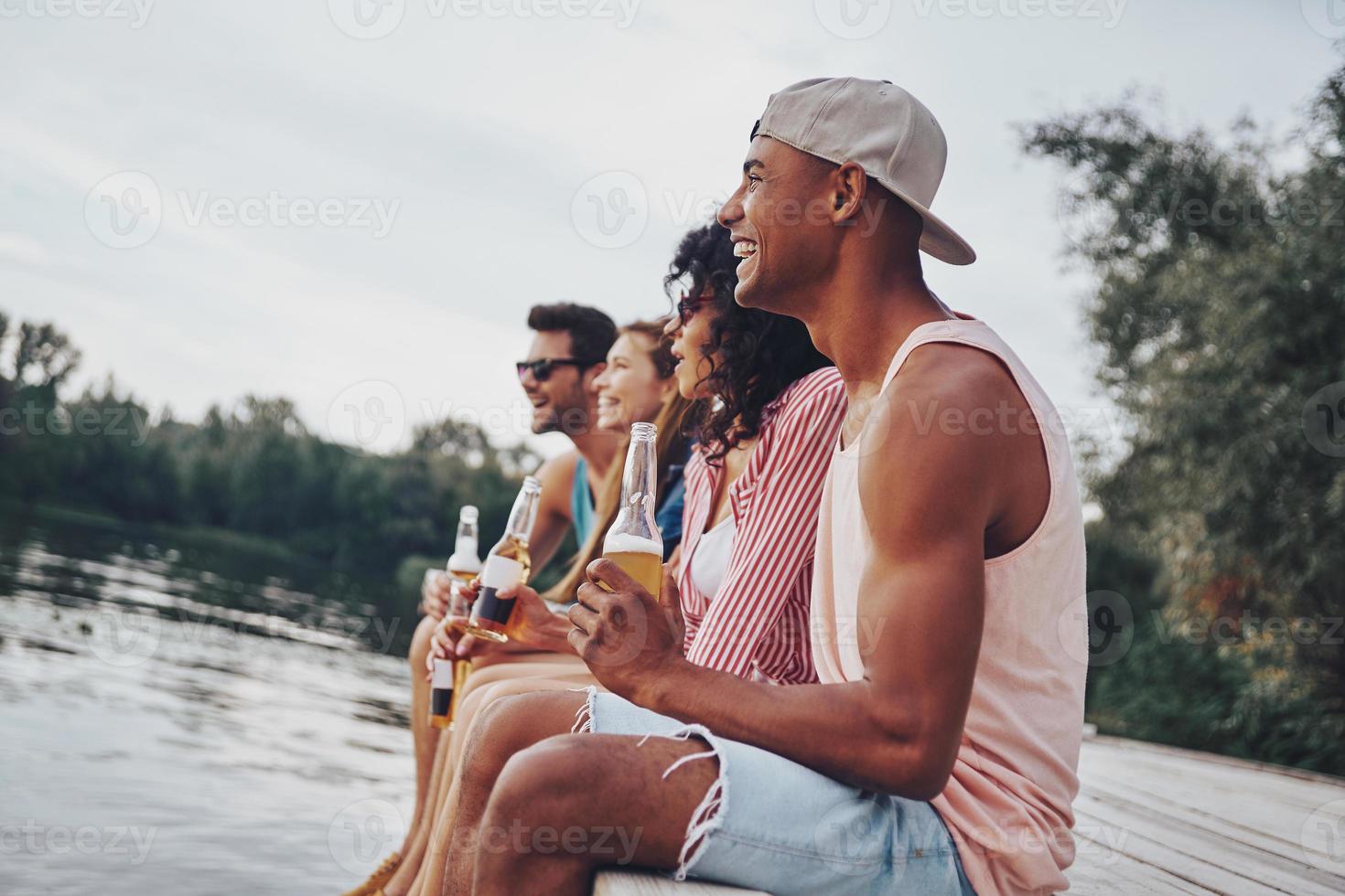 Feeling free and relaxed. Group of happy young people in casual wear smiling and drinking beer while sitting on the pier photo