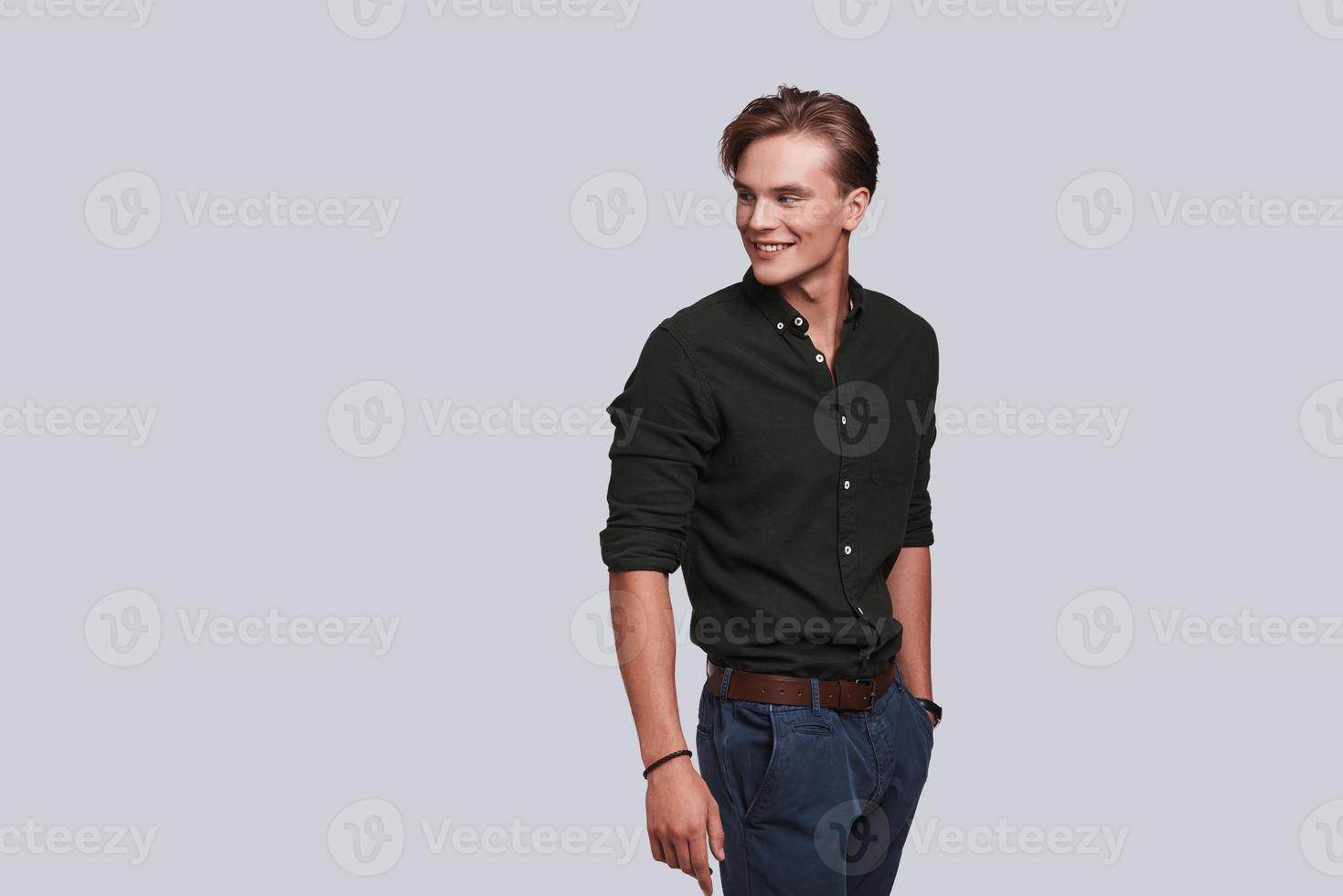 Feeling comfortable in his style. Good looking young man keeping hand in pocket and smiling while standing against grey background photo