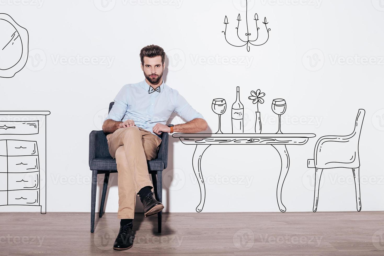 Table for two. Young handsome man keeping legs crossed and looking at camera while sitting in the chair against illustration of dining table in the background photo