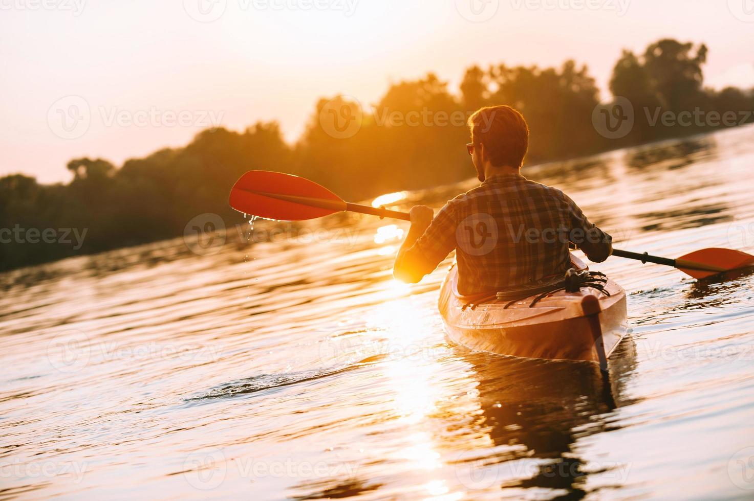 Enjoying sunset on river. Rear view of young man kayaking on lake with sunset in the background photo