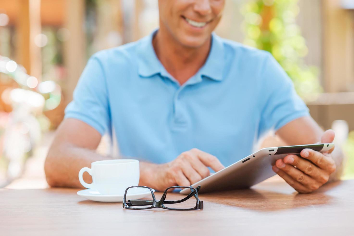 Taking advantages of free Wi-Fi. Close-up of cheerful mature man writing something in his note pad and smiling while sitting at the table outdoors with house in the background photo