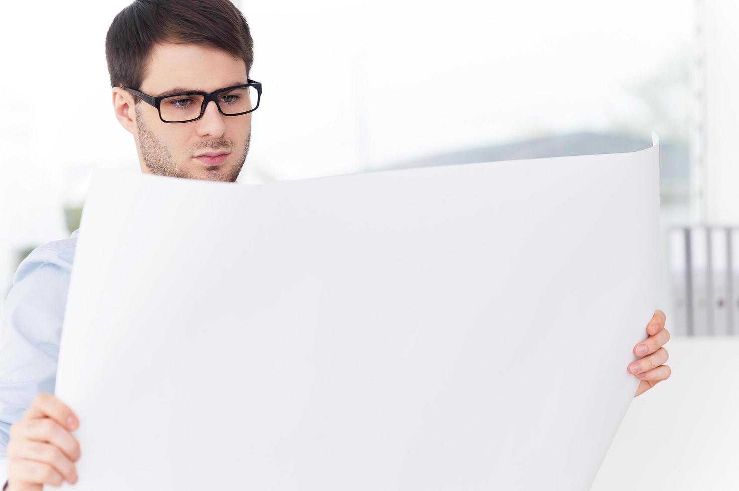 Architect with blueprint. Confident young man in shirt and tie examining blueprint while standing against the window photo