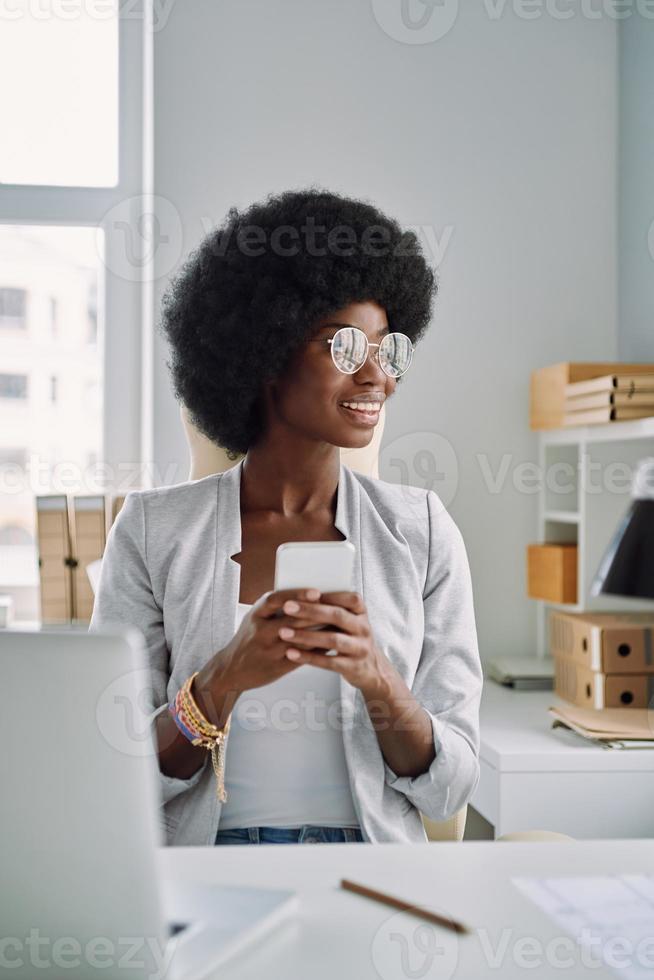 Beautiful young African woman using smart phone and smiling while sitting at her working place in office photo