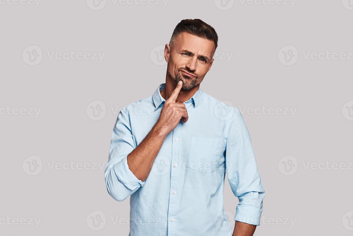 Bad idea. Thoughtful young man keeping hand on chin and making a face while standing against grey background photo