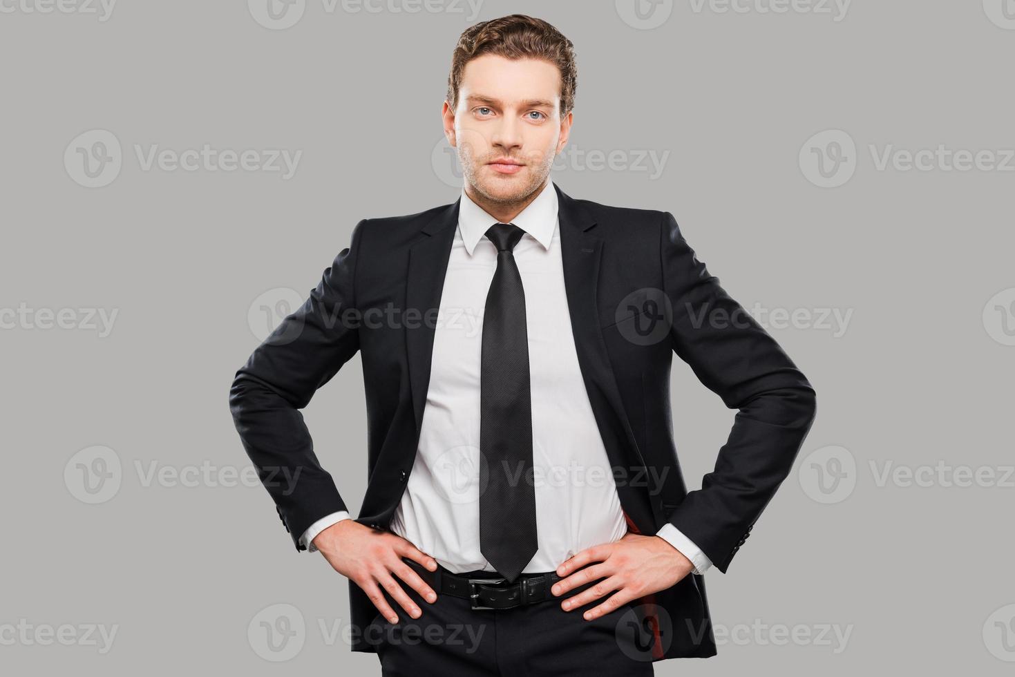 Young and successful. Confident young man in formalwear looking at camera while holding hands on hips and standing against grey background photo