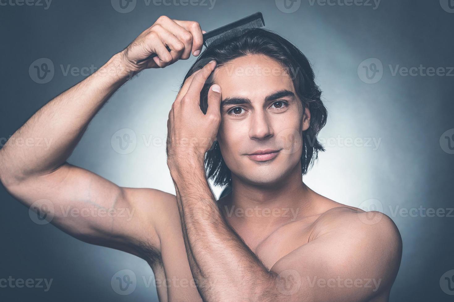 Everything should be perfect. Portrait of young shirtless man combing his hair with hairbrush and looking at camera while standing against grey background photo