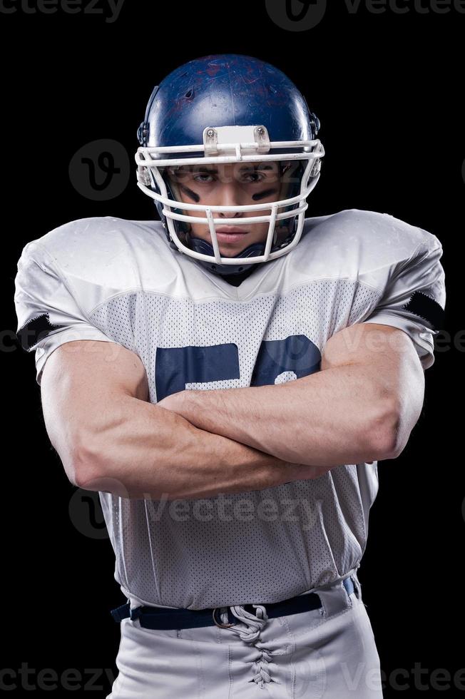 Confident in football player.  Portrait of American football player looking at camera and keeping arms crossed while standing against black background photo