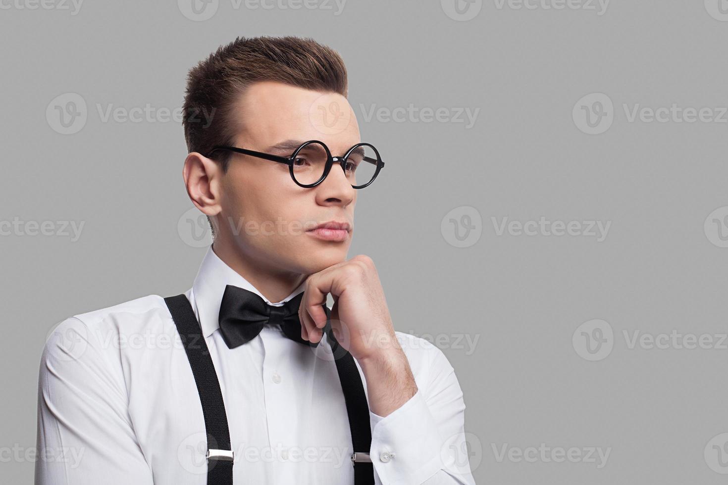 In search of inspiration. Portrait of thoughtful young nerd man in bow tie and suspenders holding hand on chin and looking at camera while standing against grey background photo